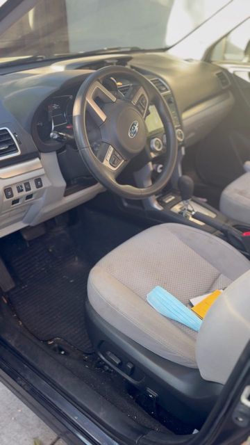Subaru driver area before cleaning, with debris on the floor mat and items on the seat